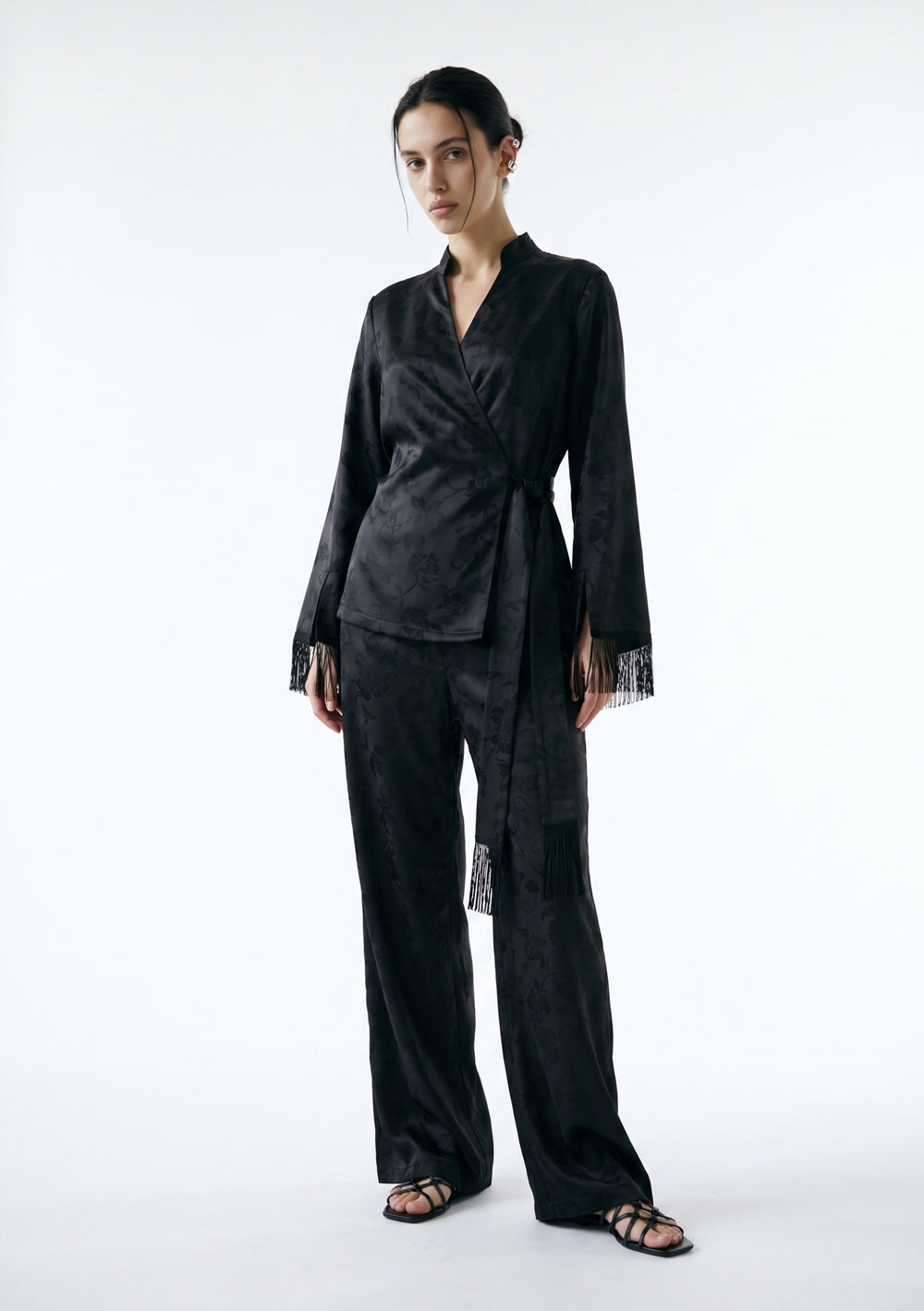 Model in the Tenebrae Set, black satin jacquard wrap top with fringed cuffs and matching wide-leg trousers, full-length three-quarter view with black strappy sandals