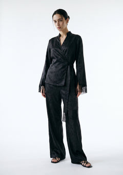 Model in the Tenebrae Set, black satin jacquard wrap top with fringed cuffs and matching wide-leg trousers, full-length three-quarter view with black strappy sandals