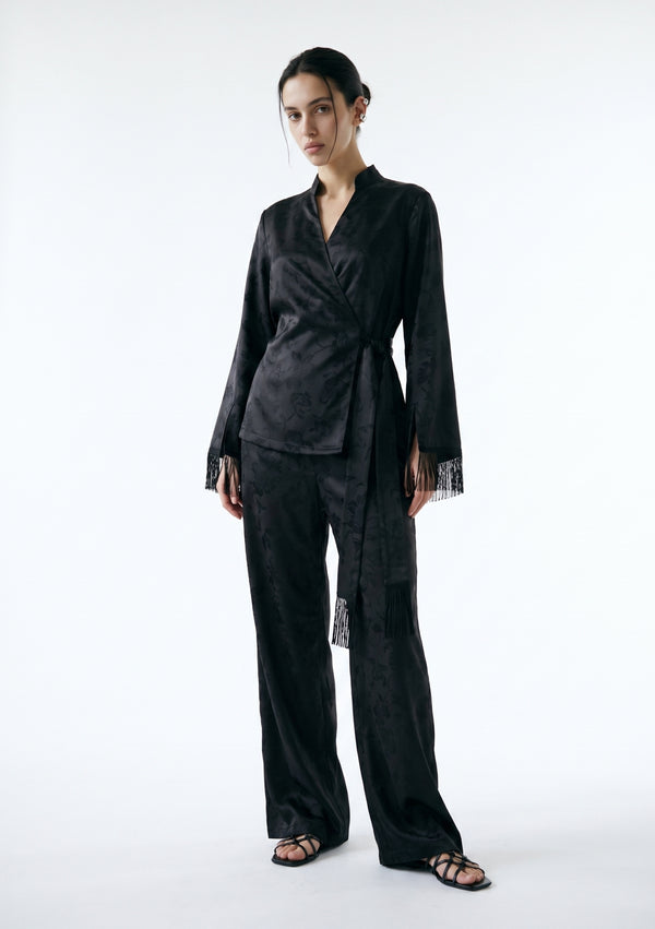 Model in the Tenebrae Set, black jacquard wrap top with fringed cuffs and matching wide-leg trousers, full-length three-quarter view with black strappy sandals