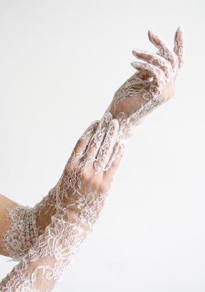 Close view of the Etherea white lace opera gloves on raised arms, showing the sheer full-fingered floral lace and eyelash-fringe cuff