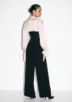 Back view of black corset trousers showing centre-back lacing, worn with a blush satin blouse.