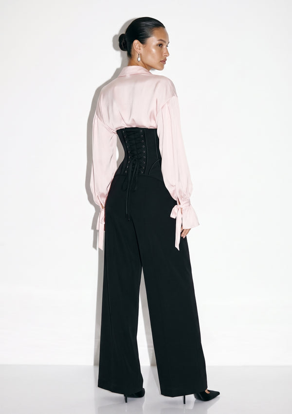 Back view of black corset trousers showing centre-back lacing, worn with a blush satin blouse.