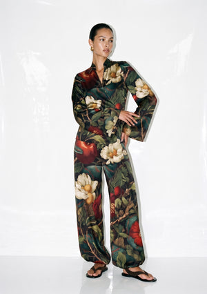 Dark green floral silk pajama set on model, wrap top with flared sleeves and wide-leg trousers in a garden print of magnolias, apples and serpents