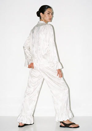 Back view of the ivory satin pajama set showing the loose shirt, wide-leg trousers and ruffled hem in movement