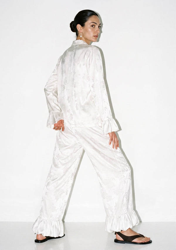 Back view of the ivory satin pajama set showing the loose shirt, wide-leg trousers and ruffled hem in movement