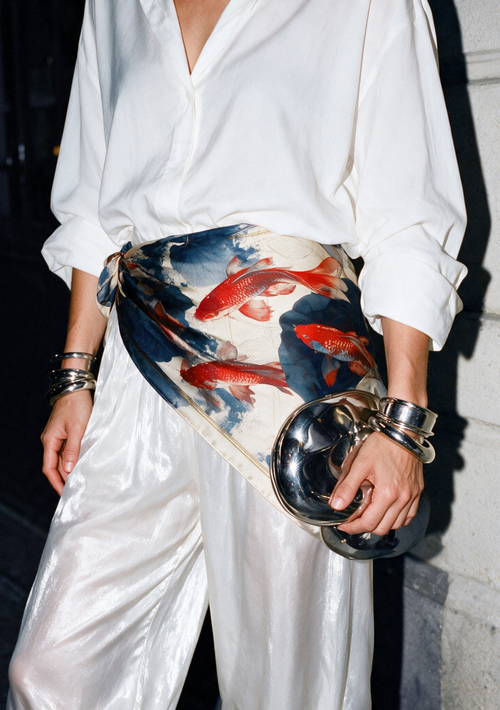 Sumi Scarf knotted as a sash at the waist over a white shirt and white satin trousers, red koi and indigo lotus silk print, worn with silver cuff bracelets