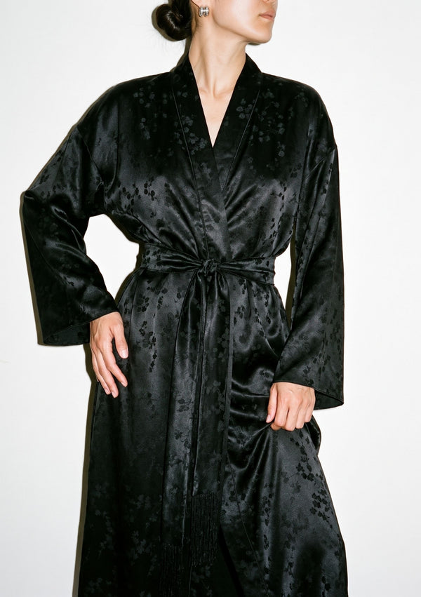 Tenebrae Robe on model lifting the hem, black satin jacquard wrap with deep V neckline and fringed belt at the waist