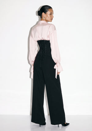 Back view of the Petal Blouse in pink viscose with full sleeves, worn with the black lace-up Elara Corset Trousers.