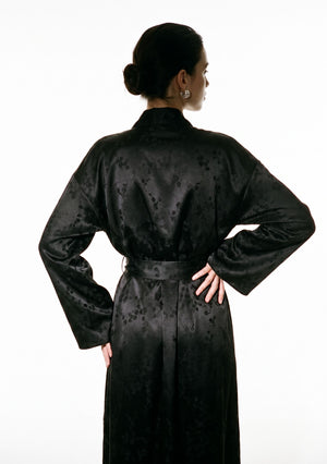 Back three-quarter view of the Tenebrae Robe, model with hands on hips, belted waist and wide sleeves in black satin jacquard
