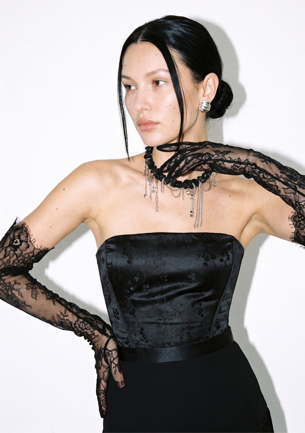 Side view of the strapless Spell Corset in black satin jacquard, with lace opera gloves and a statement necklace.