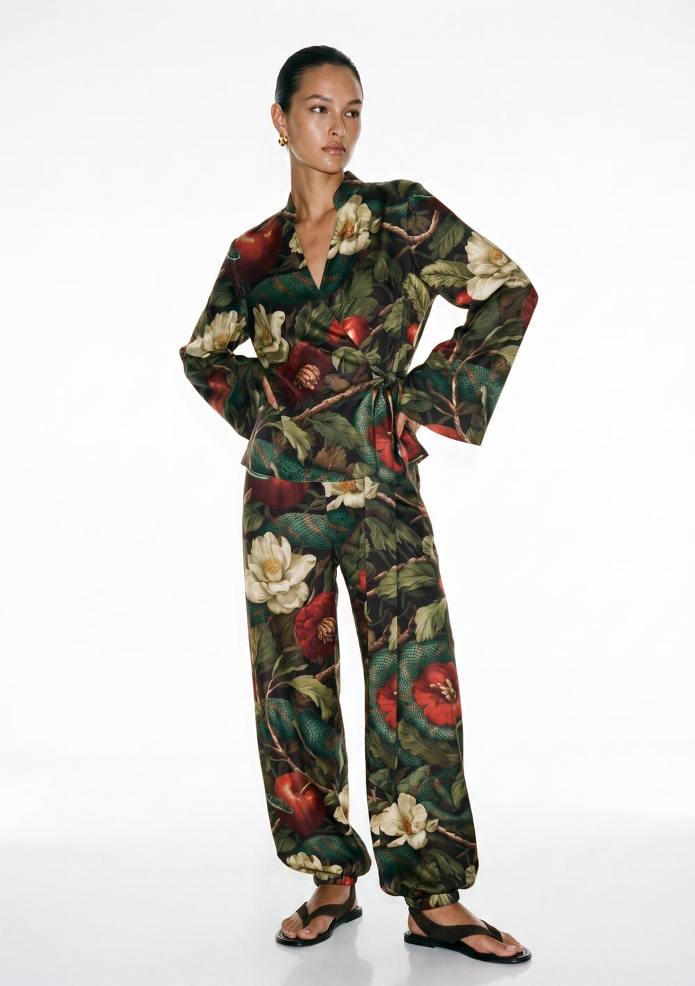 Eden silk set on model, wrap top with stand collar and wide flared cuffs tied at the waist over wide trousers gathered at the ankle, in a dark garden print of magnolias, apples and serpents